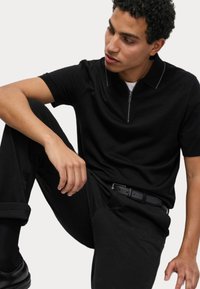 Černé polo tričko na zip s krátkým rukávem, s kontrastním límcem. Nosené s černými kalhotami a černým páskem, v sedící pozici.
