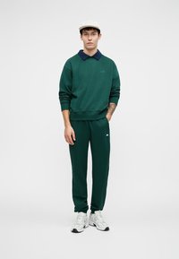 Felpa verde scuro con colletto blu navy, pantaloni verdi coordinati e scarpe bianche. L'outfit presenta una vestibilità casual e dettagli di branding minimi.
