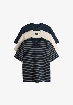 Trzy koszulki z dekoltem w łódkę nałożone warstwowo: granatowa z białymi poziomymi paskami, beżowa gładka oraz jednolita ciemnogranatowa.