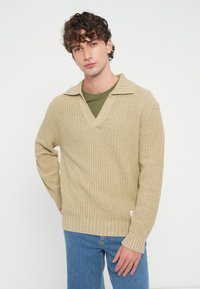 Beżowy dzianinowy sweter z dekoltem w szpic i opuszczonymi ramionami, z teksturowanym wzorem waflowym i ściągaczami przy mankietach, zestawiony z zieloną koszulą pod spodem.