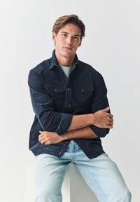 Mørkeblå manchetstribet skjorte med to brystlommer og knaplukning, kombineret med lyseblå jeans. Modellen poserer siddende med armene krydset.