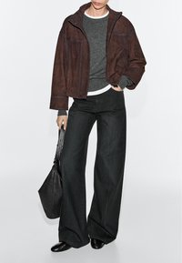 Personne portant un jean large foncé, un pull gris sous une veste marron, des bottes noires, tenant un sac à main en cuir noir sur fond blanc.