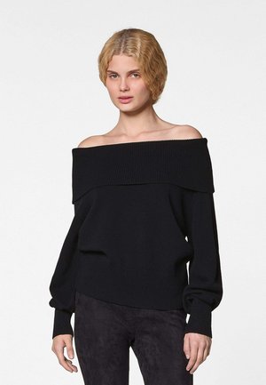 Jonge persoon met kort golvend blond haar, gekleed in een zwarte trui met blote schouders en donkere broek, staand tegen een effen achtergrond.