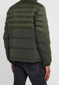 Mand i en tofarvet mørkegrøn quilted pufferjakke og sorte denimjeans, vist fra ryggen mod en ensfarvet baggrund.