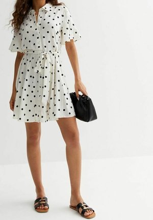 Witte dress met korte mouwen en zwarte stippen, gecincht bij de taille, met een knoopsluiting. Gecombineerd met zwarte sandalen en een bijpassende handtas.