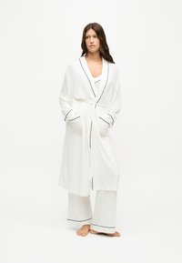 Femme debout pieds nus portant une robe blanche et un pantalon blanc ample avec un passepoil noir sur un fond blanc uni.