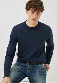 Camicia a maniche lunghe blu navy in tessuto liscio, con collo a giro e silhouette aderente, abbinata a jeans azzurri strappati.