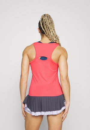 Frau mit blond geflochtenem Pferdeschwanz, die ein korallenfarbenes Racerback-Top und einen grauen und weißen, mehrlagigen Plisseerock trägt, steht mit dem Rücken zur Kamera.