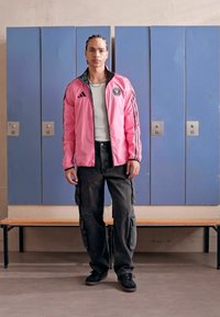 Giacca atletica leggera rosa con dettagli neri e logo, maglietta bianca a coste, pantaloni cargo neri e sneaker neri. Sfondo con armadietti.
