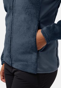 Giacca in pile di un blu navy scuro, con una texture morbida, tasca laterale per le mani con zip e pannelli in tessuto liscio sui lati e sulle maniche.