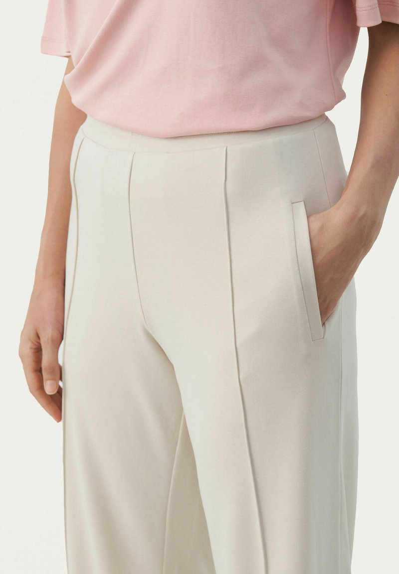 Helle beige maßgeschneiderte Hose mit Seitentaschen, sichtbaren Falten und einer glatten Textur. Rosa Kurzarmhemd, das über der Taille hängt.