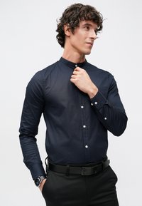 Πουκάμισο σε ναυτικό μπλε με κουμπιά μπροστά, ψηλό γιακά, εφαρμοστή γραμμή και μακριά μανίκια. Διαθέτει λευκά κουμπιά και μια μοναδική τσέπη στο στήθος.