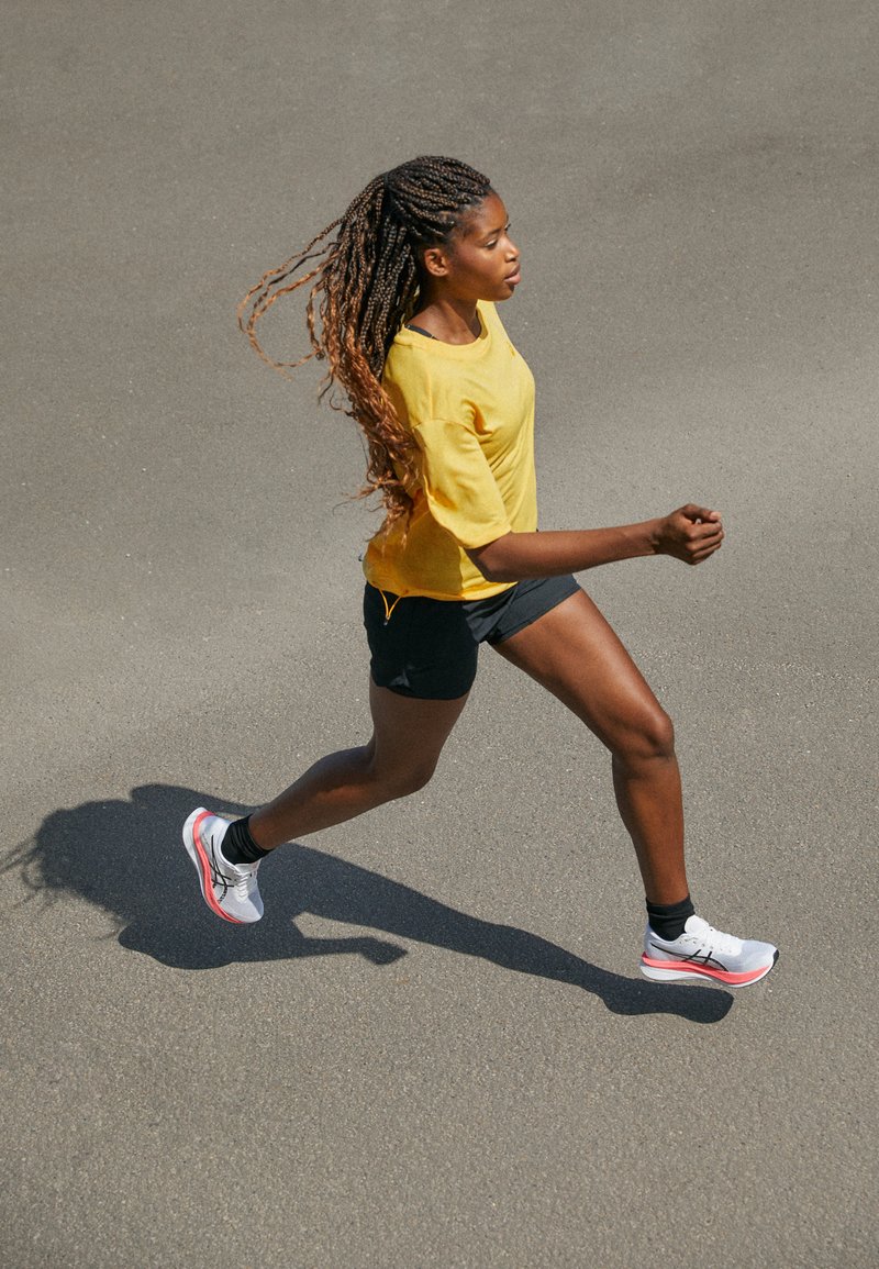Une femme court sur de l'asphalte, portant un t-shirt jaune, un short noir et des baskets blanches avec des accents roses. Ses longs cheveux tressés flottent derrière elle.