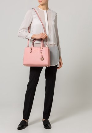 Mujer con blusa blanca y pantalones negros sosteniendo un bolso rosa Michael Kors con asa para el hombro y charm con logo dorado.