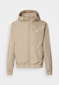 Lehká béžová větrovka s předním zipem, elastickými manžetami a kapucí, s jemným logem na levé straně hrudi.