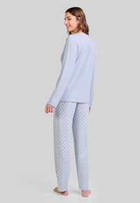 Mujer sonriendo, de pie descalza, vistiendo una blusa de manga larga azul claro y pantalones de pijama con estampado, de espaldas frente a un fondo liso.