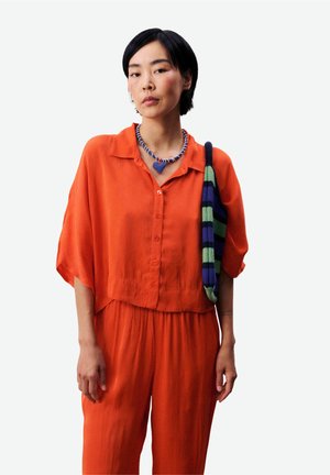 Femme aux cheveux noirs courts portant un haut à boutons orange et un pantalon, un collier de perles bleu et blanc, et un sac à bandoulière rayé bleu-vert.