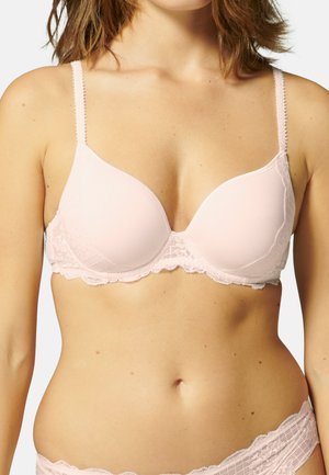 Kobieta ubrana w jasnoróżowy koronkowy biustonosz i dopasowane koronkowe majtki, pokazana od ramion do bioder na białym tle.