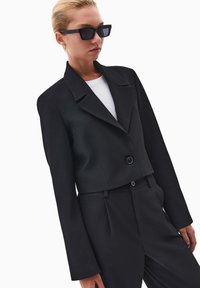 Blazer nero corto con rever a punta, chiusura con un bottone e maniche lunghe. Indossato sopra una t-shirt bianca, abbinato a pantaloni neri.