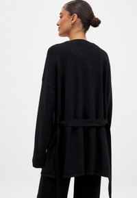 Juodas ilgomis rankovėmis megztinis su laisvu siluetu, surištomis juosmeniu ir minkštu paviršiumi. Yra apvalus nugaros hemas ir minimalūs siūlai.