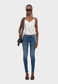 Fehér ujjatlan felső V-nyakkal, kék skinny farmer, fekete táska nagy fém részletekkel és pántos fekete szandál. A modellen napszemüveg van.