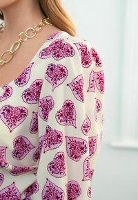 Blusa crema con motivo a cuori rosa e dettagli floreali. Maniche a sbuffo e scollo rotondo. Collana larga in oro a catena come accentuazione.