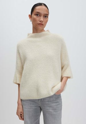 Pull en maille crème clair avec un col haut et des manches larges, présentant une texture douce. Associé à un jean gris, le modèle se tient avec les mains dans les poches.