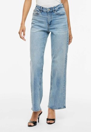 Kobieta w jasnoniebieskich jeansach z wysokim stanem i prostymi nogawkami oraz czarnych sandałach na wysokim obcasie z odkrytymi palcami, stojąca na białym tle.