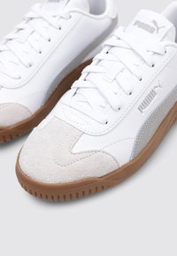 Zapatillas deportivas blancas con puntera de ante, detalles en plata y suela de goma marrón. Diseñadas con cordones planos y un acabado texturizado.