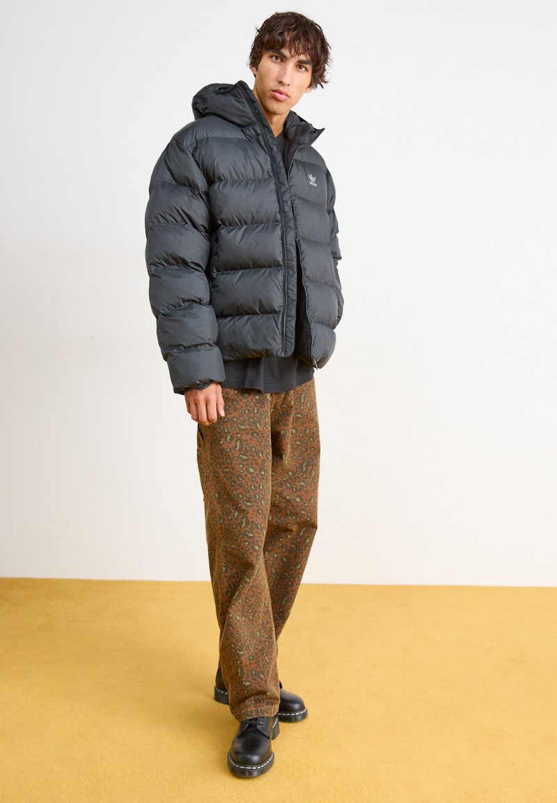 Schwarze Pufferjacke mit Kapuze, gestepptes Design, getragen über einem schwarzen Oberteil, kombiniert mit gemusterten braunen Hosen und schwarzen Schuhen.