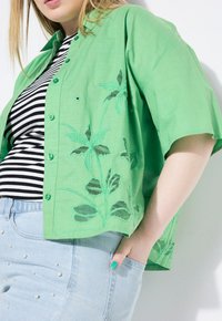 Camicia verde chiaro a maniche corte con ricami floreali, caratterizzata da una chiusura a bottoni e una vestibilità rilassata. Top a righe sottostante, jeans blu chiaro.