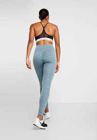 Donna che si allontana indossando un reggiseno sportivo nero, leggings azzurri chiari e scarpe da ginnastica Nike bianche su uno sfondo neutro.