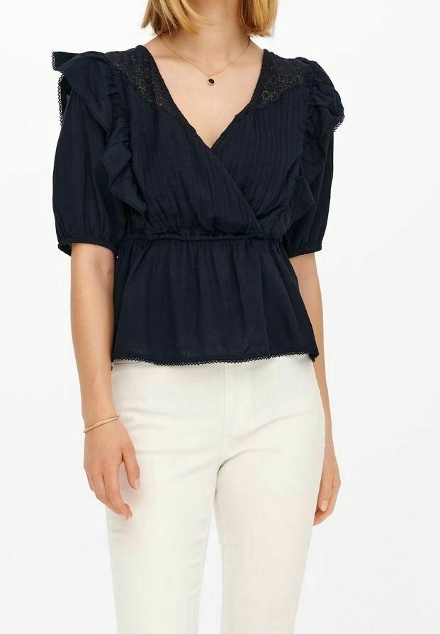 Femme portant un chemisier bleu marine à volants et manches courtes avec des détails en dentelle, associé à un pantalon blanc taille haute et un bracelet doré.