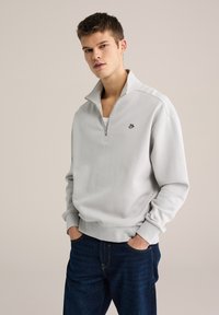 Pulover gri deschis cu guler cu fermoar pe jumătate, manșete dantelate și un logo negru mic pe piept, purtat peste o cămașă albă. Blugi din denim închis la culoare.