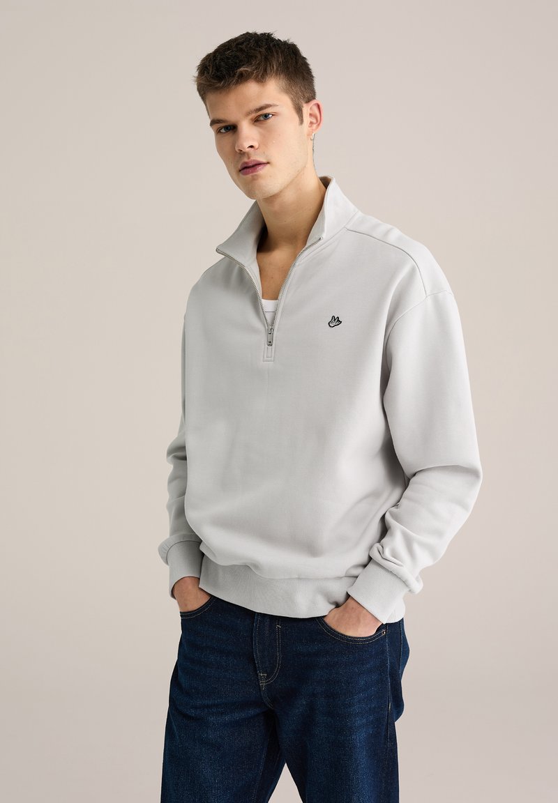 Pulover gri deschis cu guler cu fermoar pe jumătate, manșete dantelate și un logo negru mic pe piept, purtat peste o cămașă albă. Blugi din denim închis la culoare.