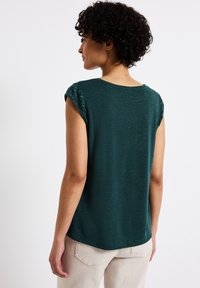 Groene top met korte mouwen van een zachte textuur, voorzien van een ronde halslijn en gaatjesdetail op de mouwen, gedragen met lichtgekleurde broek.
