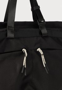Sort stof tote med justerbare stropper. Har to snørebånd med hvide og sorte accenter samt en tekstureret overflade. Holdbart design.