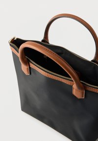 Černá taška typu tote s hnědými koženými držadly a lemováním. Má zipové zavírání se zlatým kováním. Hladká textura, otevřený vrchní design.