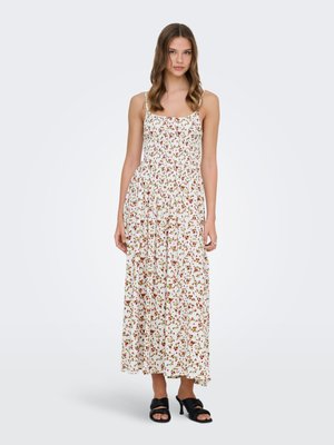 Jeune femme aux cheveux ondulés et lâchés, portant une robe maxi blanche à fleurs sans manches et des sandales noires à talons ouverts, debout sur un fond blanc.