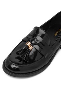 Czarne lakierowane loafersy ze szwem w podeszwie i dekoracyjnymi czarnymi frędzlami z złotymi metalowymi akcentami na białym tle.