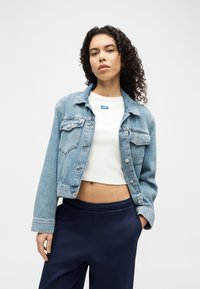 Γυναίκα με σγουρά μαλλιά που φοράει ανοιχτό μπλε τζιν μπουφάν, λευκό κοντό μπλουζάκι και μπλε σκούρο παντελόνι, στέκεται μπροστά σε απλό φόντο.