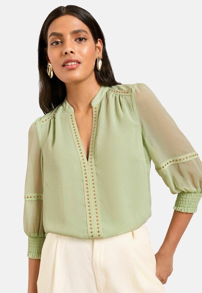 Blusa verde chiaro con scollatura a V, dettagli con bottoni dorati e maniche a sbuffo trasparenti. Presenta polsini arricciati e una texture liscia.