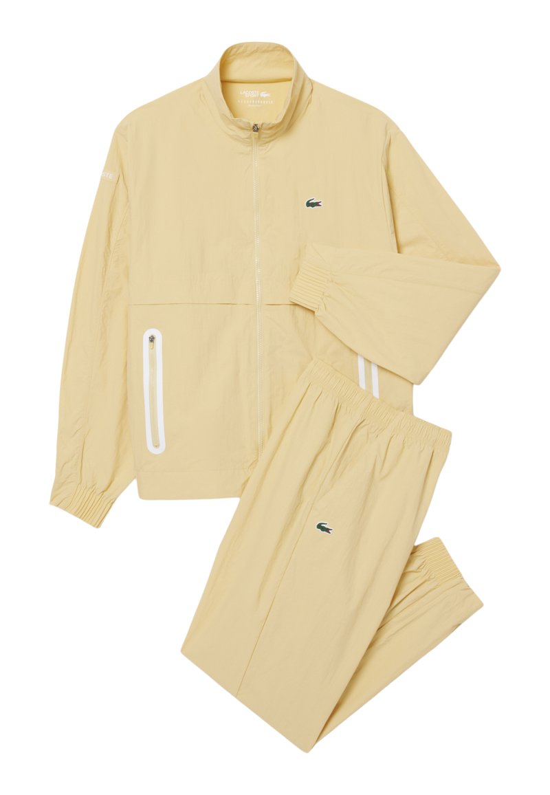 Beige trainingspak met ritsjack en broeken met elastische taille, met kleine groene krokodillenlogo's op jas en broeken.