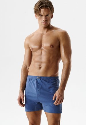 Mężczyzna o umięśnionej sylwetce i krótkich blond włosach, ubrany w niebieskie bokserki, stoi na tle jednolitego białego tła.