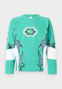 Πράσινο αθλητικό πουκάμισο μακρυμάνικο με μαύρα και λευκά αφηρημένα σχέδια και κεντρικό κίτρινο λογότυπο Puma μέσα σε μαύρο και λευκό οβάλ με αιχμές.