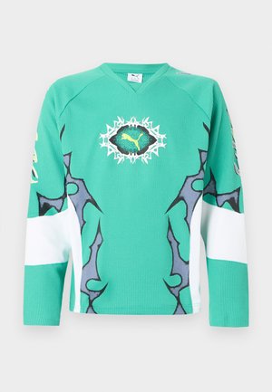 Grünes Sporttrikot mit langen Ärmeln, schwarzen und weißen abstrakten Mustern und einem zentralen gelben Puma-Logo innerhalb eines schwarz-weiß gezackten Ovals.