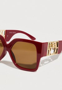 Κόκκινα ορθογώνια γυαλιά ηλίου με καφέ φακούς. Διαθέτουν γυαλιστερή επιφάνεια και χρυσά μεταλλικά στοιχεία με ξεχωριστό λογότυπο στους βραχίονες.