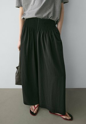 Person trägt lockere schwarze, weit geschnittene Hosen, graues kurzärmeliges Hemd, rote Flip-Flops und hält eine braune Handtasche vor einem schlichten Hintergrund.