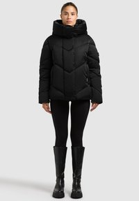 Czarna kurtka puchowa z wysokim kołnierzem, ozdobiona pikowanym, geometrycznym wzorem. Zestawiona z czarnymi legginsami i kozakami za kolano.