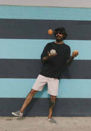 Mężczyzna w okularach przeciwsłonecznych, czarnej koszulce i szortach żongluje trzema piłkami na tle niebiesko-czarnej pasiasty ściany, balansując na jednej nodze.